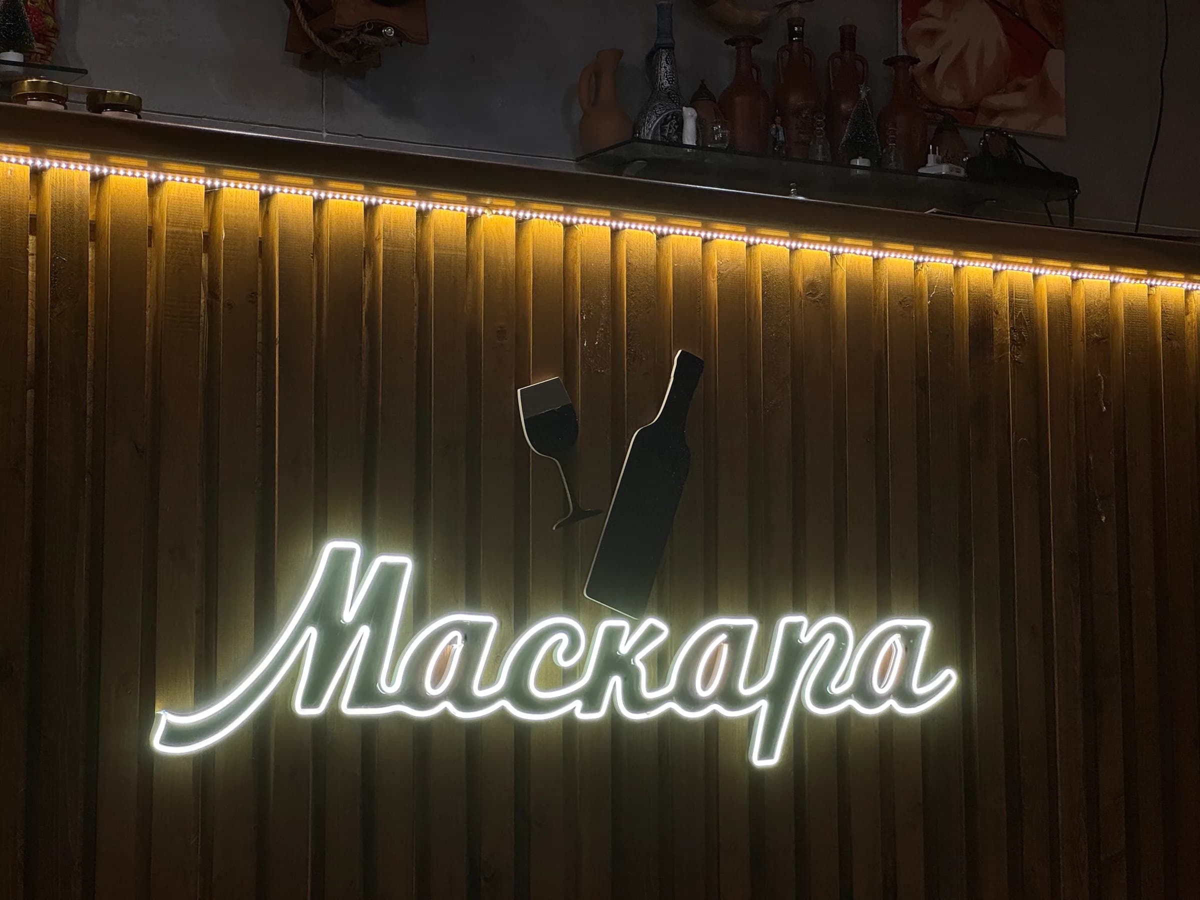 Неоновая вывеска «Маскара» с бокалом и бутылкой на реечной стене бара, тёплый жёлтый свет